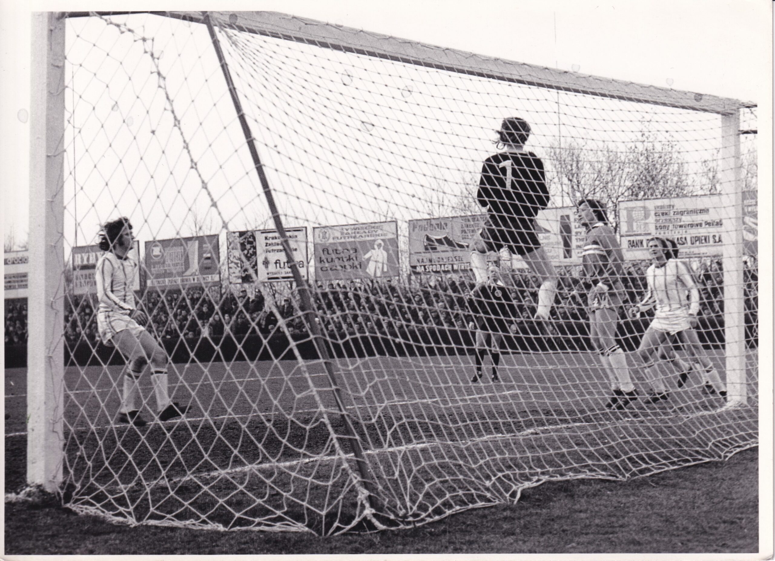 Mecz 1/4 finału Pucharu Polski Garbarnia Kraków - Ruch Chorzów rozegrany w marcu 1974 na stadionie Korony przy ul. Parkowej w Krakowie, Bramkarz Garbarni Tadeusz Draus w wyskoku do piłki, po jego prawej stronie obrońca Garbarni Marek Piwowarczyk. W polu karnym Garbarni stoi dwóch zawodników Ruchu i sędzia Karolak. W tle wypełniona widzami północna trybuna z tablicami reklamowymi.